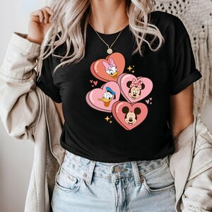 Puede incluir: Camiseta negra con un gr&aacute;fico de Mickey Mouse, Minnie Mouse, Donald Duck y Daisy Duck en corazones rosas.