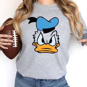 May include: A gray sweatshirt featuring a cartoon image of Donald Duck's face. The design includes a blue sailor hat, a yellow beak, and an angry expression. The person in the image is holding a brown football.
