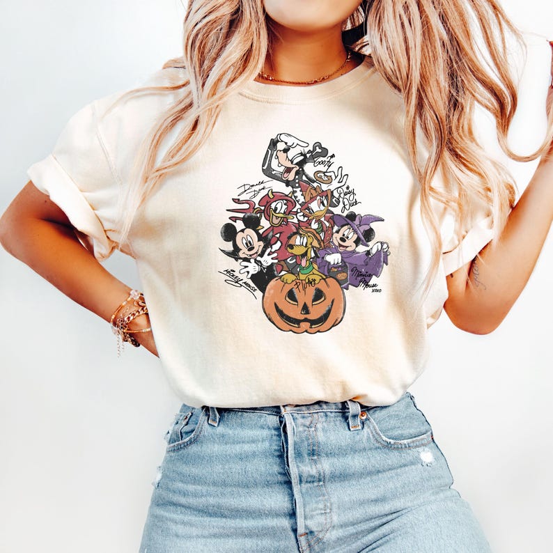 May include: Cream-colored t-shirt featuring a Halloween-themed graphic. The graphic includes various cartoon characters, including Mickey Mouse, Donald Duck, and Goofy, surrounding a carved pumpkin. The design has a vintage, slightly faded appearance.