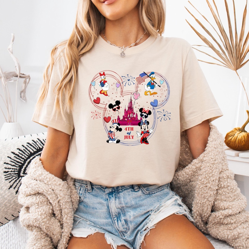 Peut inclure: T-shirt beige avec un motif color&eacute; de personnages Disney c&eacute;l&eacute;brant le 4 juillet. Le motif comprend Mickey et Minnie Mouse, Donald Duck et Daisy Duck, avec des feux d'artifice et un ch&acirc;teau. Le texte "4th of July" est &eacute;galement inclus.