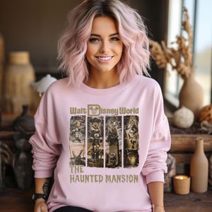May include: A pink sweatshirt featuring a graphic of the Haunted Mansion attraction at Walt Disney World. The graphic depicts the iconic characters from the ride, including the Ghost Host, Constance Hatchaway, and the Hitchhiking Ghosts. The text "Walt Disney World" and "The Haunted Mansion" is printed in gold.