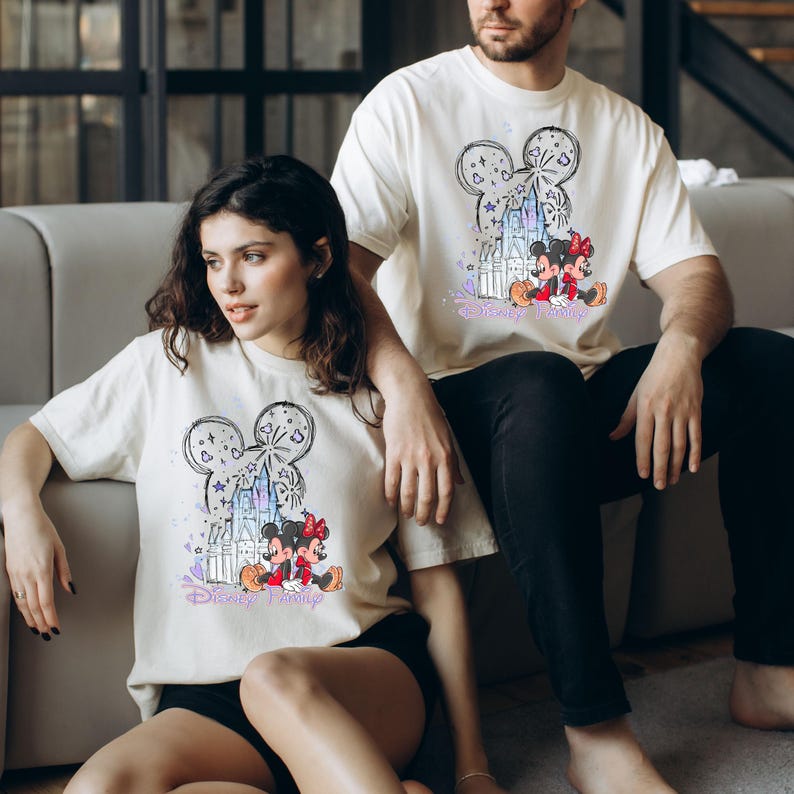 May include: Off-white t-shirts with a Mickey Mouse head design, a castle, fireworks, and the words "Disney Family." The graphic includes Mickey and Minnie Mouse in red and black. The shirts are worn by two people.
