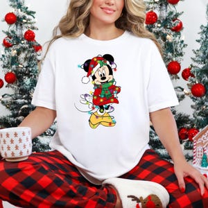 K&ouml;nnte beinhalten: Wei&szlig;es T-Shirt mit einem weihnachtlichen Minnie Mouse-Motiv. Minnie Mouse ist in einem festlichen Outfit dargestellt, einschlie&szlig;lich rotem Mantel, gr&uuml;nem Schal und Weihnachtsm&uuml;tze, alles mit Lichterketten geschm&uuml;ckt.