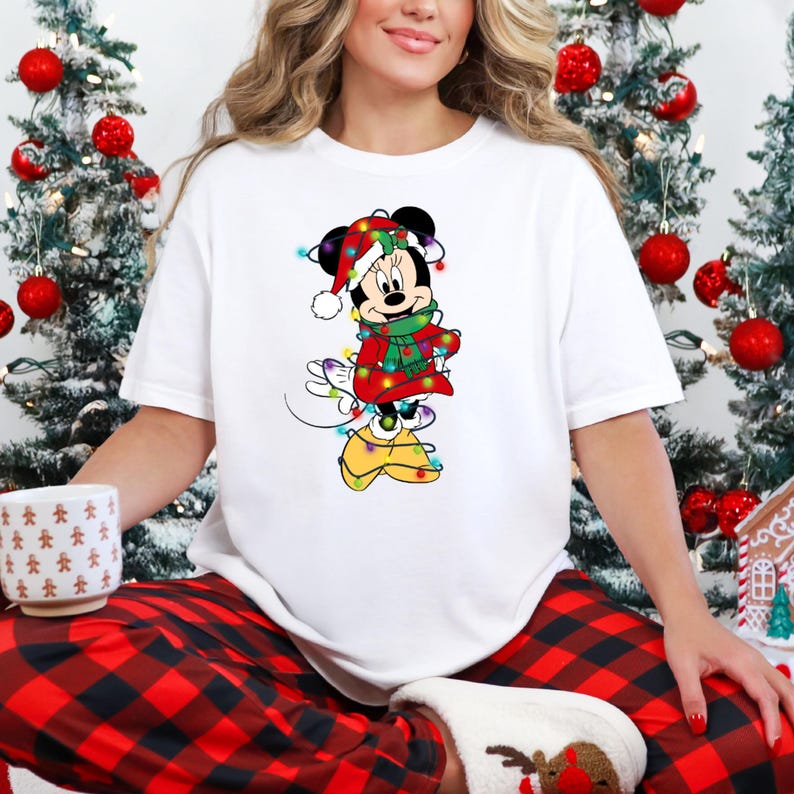 K&ouml;nnte beinhalten: Wei&szlig;es T-Shirt mit einem weihnachtlichen Minnie Mouse-Motiv. Minnie Mouse ist in einem festlichen Outfit dargestellt, einschlie&szlig;lich rotem Mantel, gr&uuml;nem Schal und Weihnachtsm&uuml;tze, alles mit Lichterketten geschm&uuml;ckt.