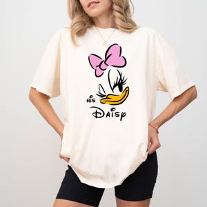 May include: Cream-colored t-shirt featuring a graphic of Daisy Duck with a pink bow, black eyelashes, and a yellow beak. The text "His Daisy" is printed below the character in black script. The shirt is a casual style.