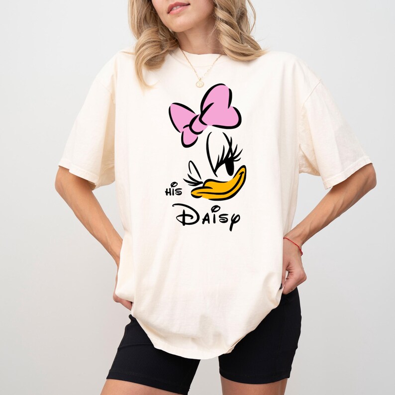 May include: Cream-colored t-shirt featuring a graphic of Daisy Duck with a pink bow, black eyelashes, and a yellow beak. The text "His Daisy" is printed below the character in black script. The shirt is a casual style.