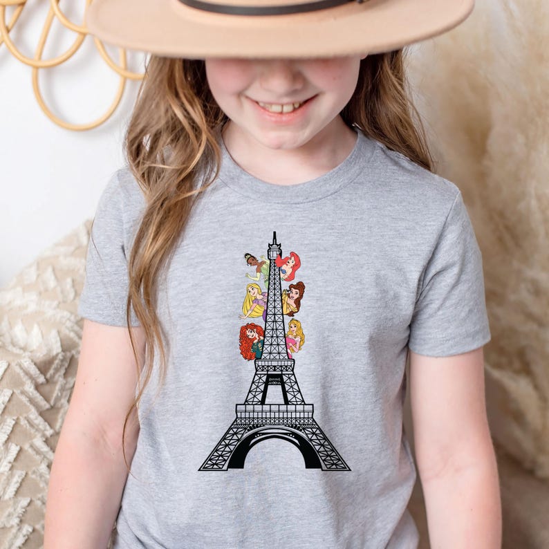 May include: A gray t-shirt featuring a graphic of the Eiffel Tower with several cartoon princesses. The Eiffel Tower is black and white, and the princesses are in various colors. The shirt is being worn by a person.
