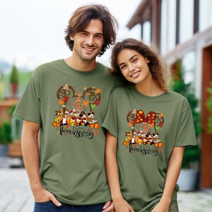 May include: Two olive green t-shirts featuring a Thanksgiving design. The design includes cartoon characters, pumpkins, and the words "Happy Thanksgiving" in a script font. The shirts are worn by a man and a woman.