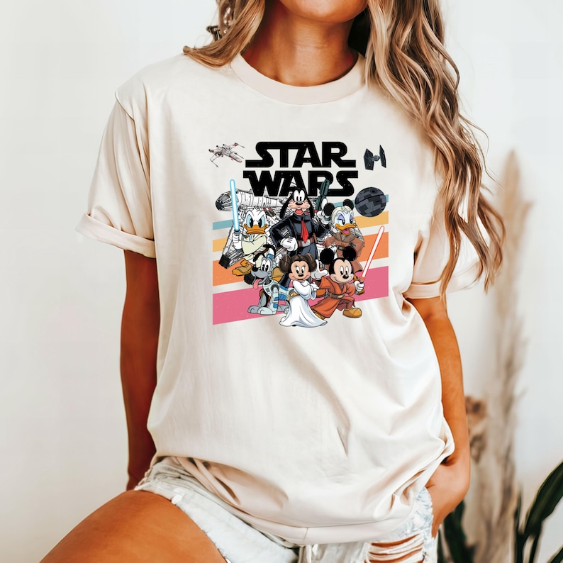 K&ouml;nnte beinhalten: Beiges T-Shirt mit einer farbenfrohen Grafik von Disney-Charakteren in Star Wars-Kost&uuml;men. Das Star Wars-Logo ist &uuml;ber den Charakteren aufgedruckt. Das Design umfasst Micky Maus, Minnie Maus, Donald Duck und Goofy.