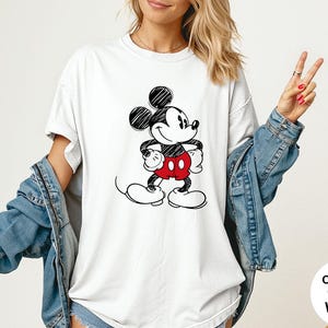 May include: White t-shirt featuring a black and red Mickey Mouse graphic. The cartoon character is drawn in a sketch style. The shirt is part of the Comfort Colors collection.