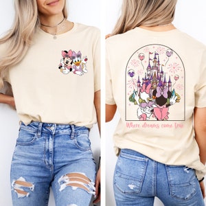 Pu&ograve; includere: Una maglietta beige chiaro con un'immagine di Topolino e Minnie Mouse sul davanti e un'immagine del castello di Cenerentola con fuochi d'artificio e il testo "Where dreams come true" sul retro.