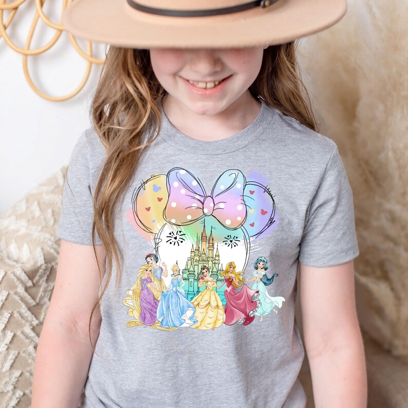 May include: A gray t-shirt with a rainbow-colored Minnie Mouse head design featuring a bow, Cinderella's castle, and Disney princesses. The princesses are wearing their signature dresses and are standing in front of the castle.