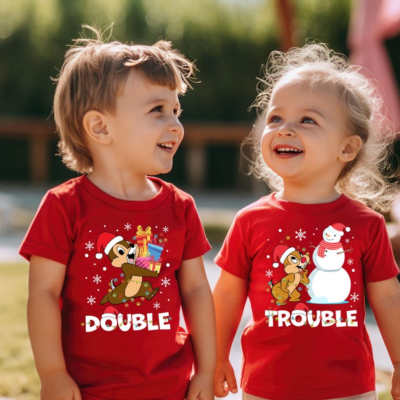 Puede incluir: Dos ni&ntilde;os con camisetas rojas de tem&aacute;tica navide&ntilde;a. La camiseta de la izquierda dice "Double" y presenta una ardilla de dibujos animados sosteniendo regalos. La camiseta de la derecha dice "Trouble" y presenta una ardilla de dibujos animados y un mu&ntilde;eco de nieve.