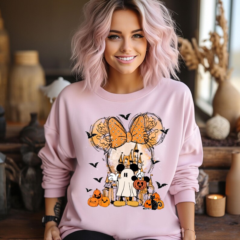 May include: Pink sweatshirt with a graphic of Mickey Mouse and Minnie Mouse dressed in Halloween costumes. Minnie Mouse is wearing a large orange bow with white polka dots and a ghost costume. Mickey Mouse is dressed as a ghost. The graphic also includes a castle, bats, and pumpkins.