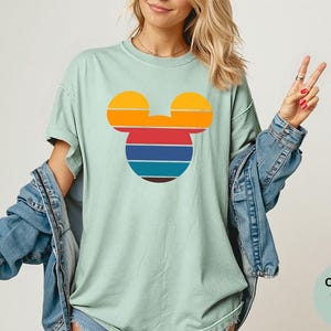 K&ouml;nnte beinhalten: Hellgr&uuml;nes T-Shirt mit einem Retro-Design eines Mickey Mouse-Kopfes. Das Design umfasst horizontale Streifen in Orange, Rot, Blau und Braun. Das Shirt ist Teil der Comfort Colors Bay-Kollektion.