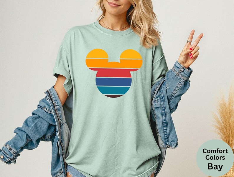 K&ouml;nnte beinhalten: Hellgr&uuml;nes T-Shirt mit einem Retro-Design eines Mickey Mouse-Kopfes. Das Design umfasst horizontale Streifen in Orange, Rot, Blau und Braun. Das Shirt ist Teil der Comfort Colors Bay-Kollektion.