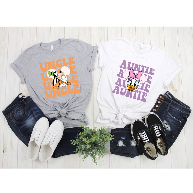 Puede incluir: Dos camisetas con dise&ntilde;os gr&aacute;ficos y texto. Una es gris con "UNCLE" en naranja y un personaje de dibujos animados. La otra es blanca con "AUNTIE" en morado y un personaje de dibujos animados. Ambas camisetas se combinan con vaqueros y zapatos.