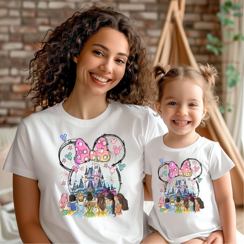 May include: A white t-shirt with a graphic of a pink bow, a castle, and fireworks. The graphic is surrounded by a black outline of Mickey Mouse ears. There are also several princesses in the graphic.
