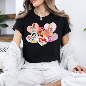 Pode incluir: Camiseta preta com um gr&aacute;fico de princesas Disney em cora&ccedil;&otilde;es. As princesas incluem Ariel, Bela, Cinderela, Branca de Neve, Tiana e Rapunzel.