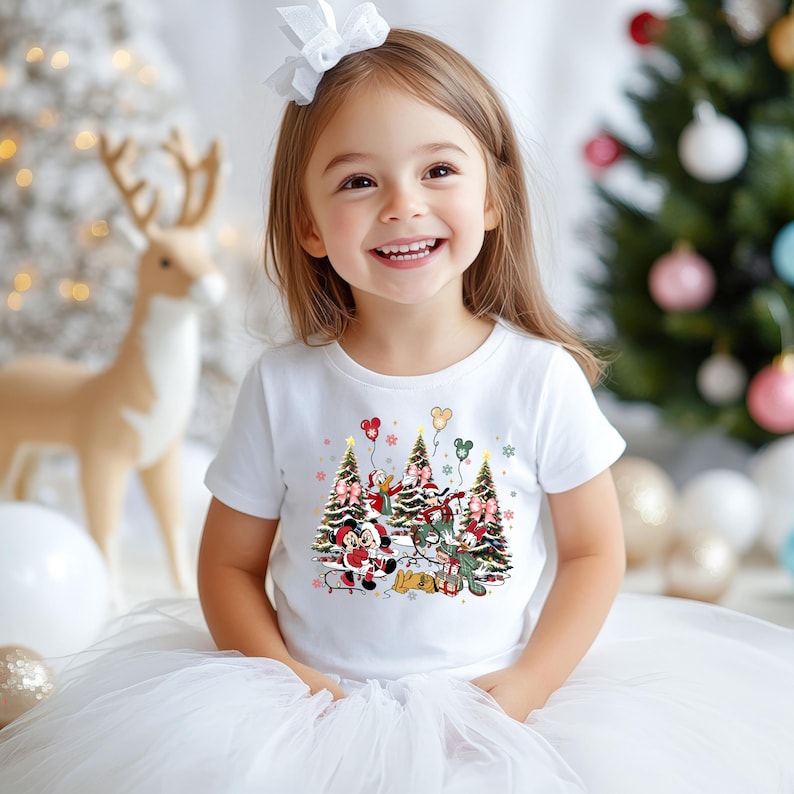 May include: White toddler t-shirt features a Christmas-themed design. Mickey Mouse, Minnie Mouse, Donald Duck, and Goofy are depicted around two decorated Christmas trees with balloons. The design is in shades of green, red, and white against a white background. The shirt is a comfortable, casual style, perfect for the holiday season.