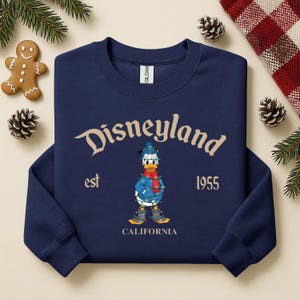 May include: Navy blue sweatshirt with "Disneyland" arched text and a graphic of Donald Duck in a winter outfit. The text "est 1955 CALIFORNIA" is also printed on the sweatshirt. The sweatshirt is surrounded by Christmas decorations.