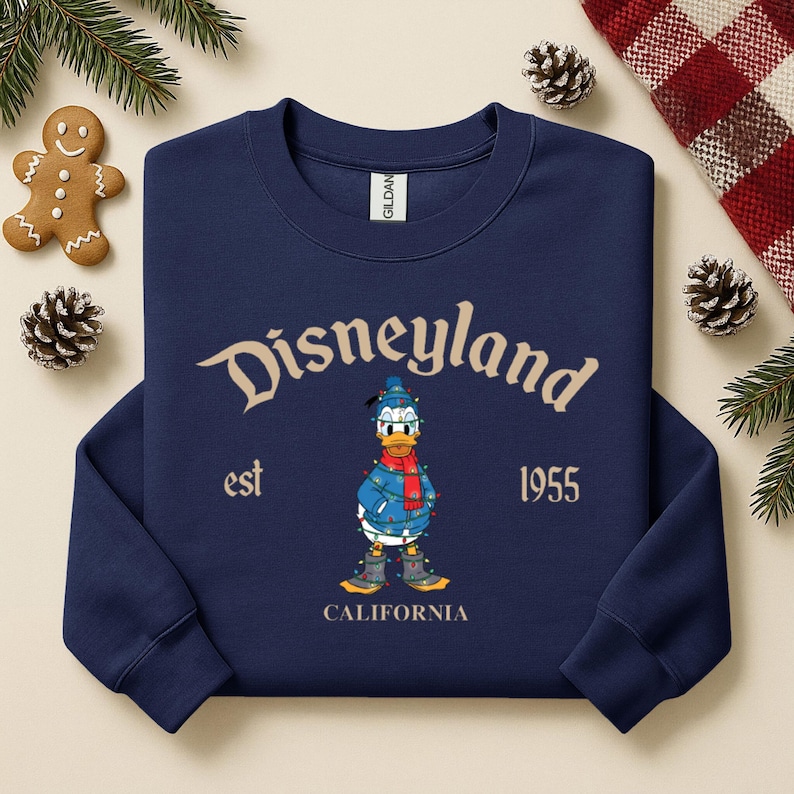May include: Navy blue sweatshirt with "Disneyland" arched text and a graphic of Donald Duck in a winter outfit. The text "est 1955 CALIFORNIA" is also printed on the sweatshirt. The sweatshirt is surrounded by Christmas decorations.