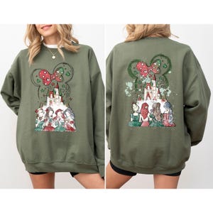 May include: Olive green sweatshirt featuring a castle graphic with a Minnie Mouse bow and fireworks. The front and back of the sweatshirt display a castle and princess design. The design includes red, white, and green accents.