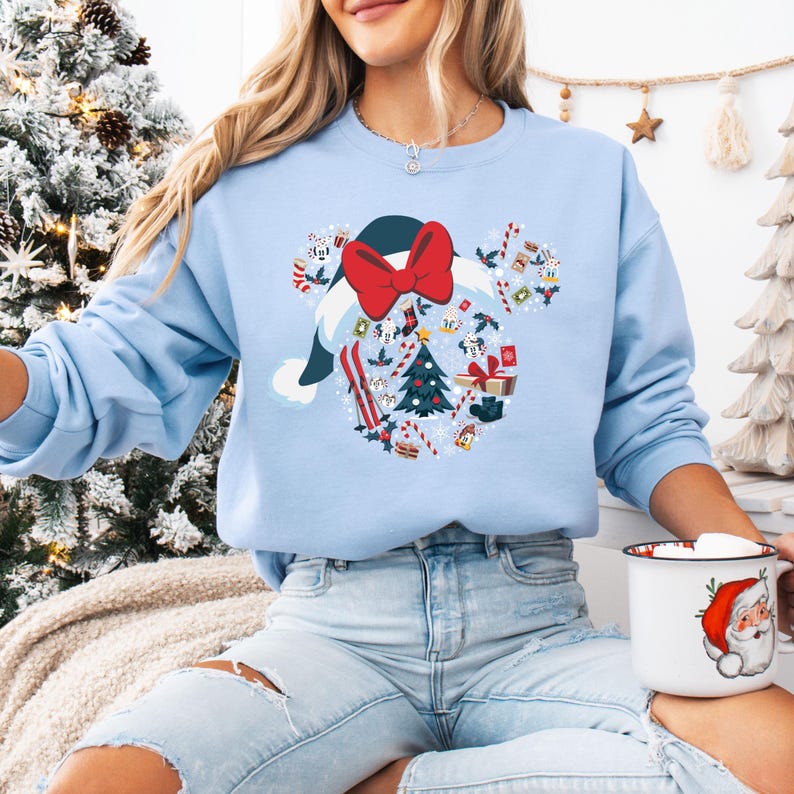 May include: Pale blue sweatshirt with a festive design. The graphic is shaped like Mickey Mouse ears, incorporating a Santa hat, red bow, Christmas tree, skis, and gift boxes. The sweatshirt has long sleeves and a crew neck.