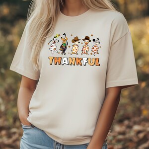 May include: Beige t-shirt featuring a graphic of cartoon dogs in festive attire, with the word "THANKFUL" in orange and brown block letters. The dogs are decorated with fall leaves and hats, creating a Thanksgiving theme.