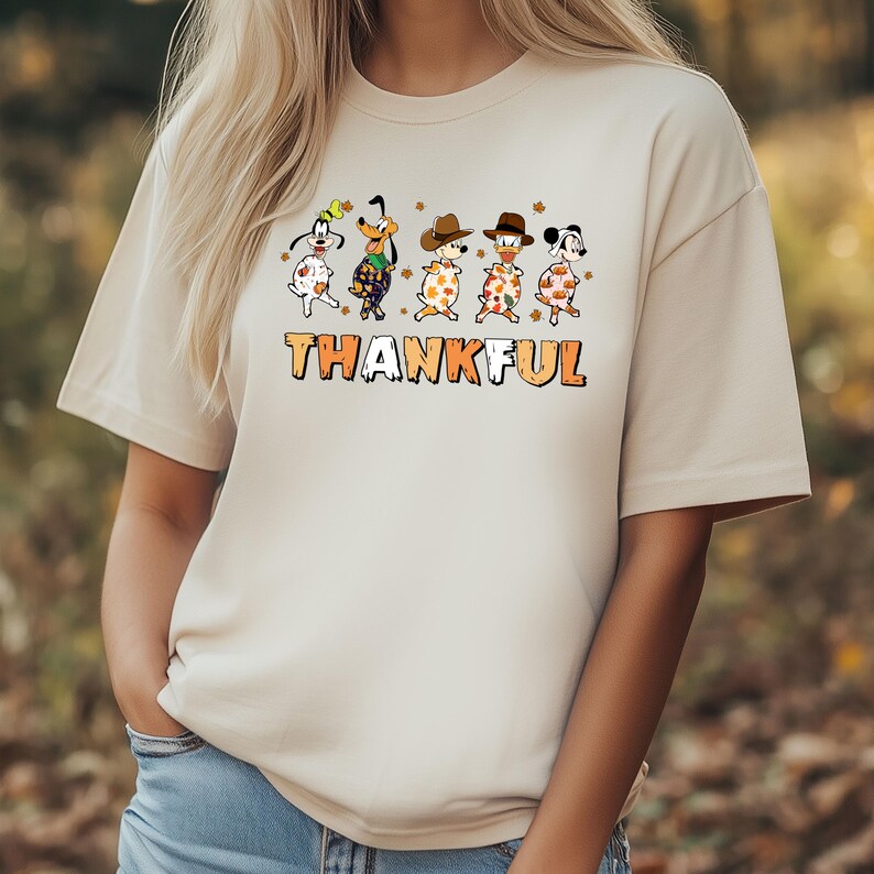 May include: Beige t-shirt featuring a graphic of cartoon dogs in festive attire, with the word "THANKFUL" in orange and brown block letters. The dogs are decorated with fall leaves and hats, creating a Thanksgiving theme.