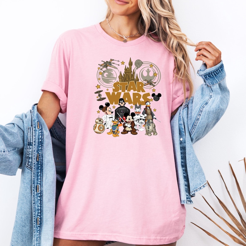 May include: A light pink t-shirt featuring a Star Wars graphic with Disney characters. The design includes the Star Wars logo, a castle, spaceships, and cartoon characters like Mickey Mouse and Donald Duck.