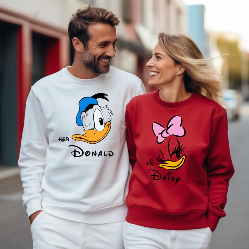 May include: A couple wearing matching sweatshirts. One sweatshirt is white with a Donald Duck graphic and the text "Her Donald". The other is red with a Daisy Duck graphic and the text "His Daisy".