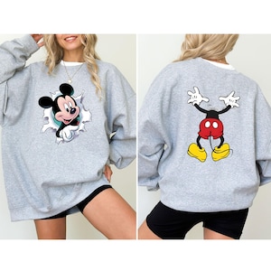 Puede incluir: Una sudadera gris claro con un dise&ntilde;o de Mickey Mouse en la parte delantera y trasera. El gr&aacute;fico frontal muestra a Mickey rompiendo un efecto de papel rasgado. El gr&aacute;fico trasero representa las piernas y los brazos de Mickey. La sudadera tiene un cuello blanco.