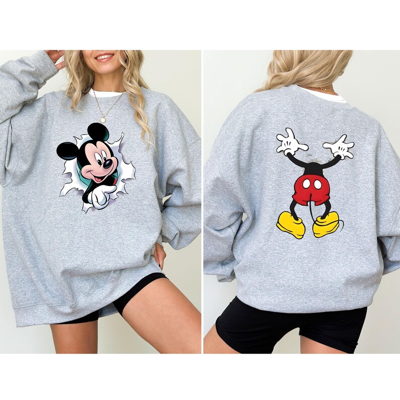 Puede incluir: Una sudadera gris claro con un dise&ntilde;o de Mickey Mouse en la parte delantera y trasera. El gr&aacute;fico frontal muestra a Mickey rompiendo un efecto de papel rasgado. El gr&aacute;fico trasero representa las piernas y los brazos de Mickey. La sudadera tiene un cuello blanco.