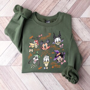 Puede incluir: Una sudadera verde oliva con un gr&aacute;fico colorido de personajes de Disney, incluyendo Mickey Mouse, Minnie Mouse, Donald Duck y Pluto. El dise&ntilde;o incluye murci&eacute;lagos, lazos y estrellas, con las palabras "Mickey Mouse" y "Minnie Mouse".