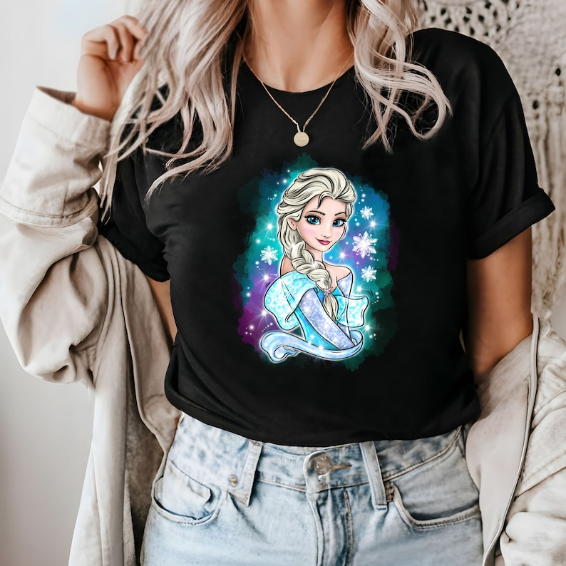 Op de afbeelding: Een zwart T-shirt met een kleurrijke illustratie van een cartoonprinses met lang blond haar en een blauwe jurk. De prinses wordt omringd door sneeuwvlokken en sterren.