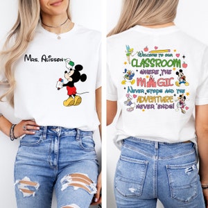 Puede incluir: Camiseta blanca con una ilustraci&oacute;n de Mickey Mouse sosteniendo un l&aacute;piz. La parte posterior de la camiseta tiene un dise&ntilde;o colorido con el texto "Bienvenido a nuestra aula donde la magia nunca se detiene y la aventura nunca termina!"