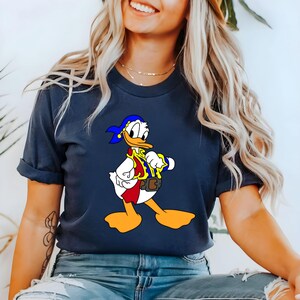 May include: Navy blue t-shirt featuring a cartoon image of Donald Duck dressed as a pirate. The graphic includes a blue bandana, red vest, and yellow accents. The shirt is a casual style.