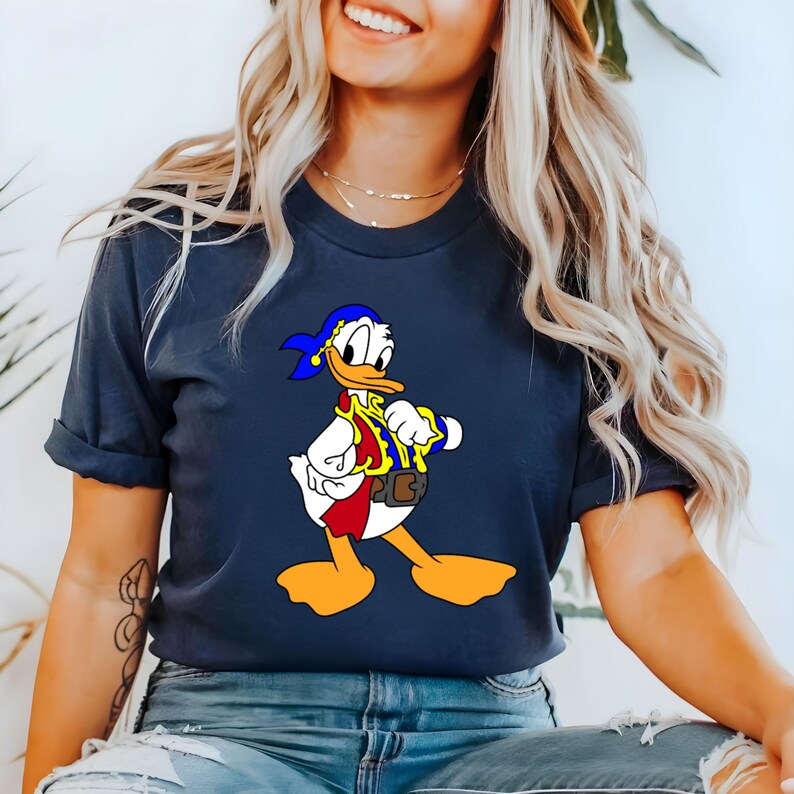 May include: Navy blue t-shirt featuring a cartoon image of Donald Duck dressed as a pirate. The graphic includes a blue bandana, red vest, and yellow accents. The shirt is a casual style.