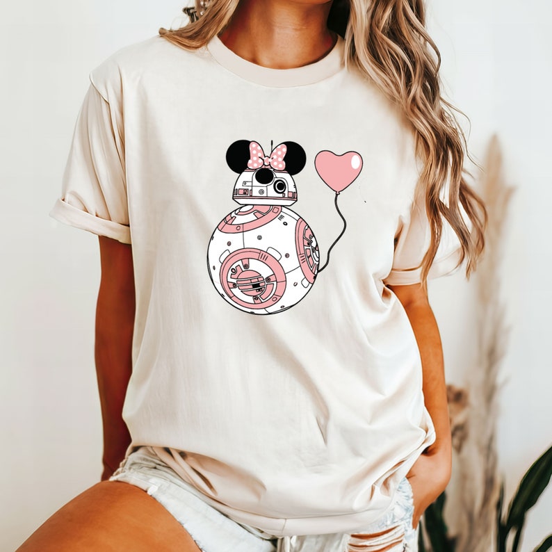 May include: Cream-colored t-shirt featuring a whimsical design. The graphic depicts a droid-like character with Minnie Mouse ears and a pink bow, holding a pink heart-shaped balloon. The droid is white with pink accents.