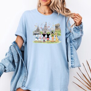 May include: Light blue t-shirt featuring a colorful illustration of Disney characters, including Mickey and Minnie Mouse, Donald and Daisy Duck, Goofy, and Pluto, in front of a castle and fireworks display. The shirt is a comfortable, casual style.
