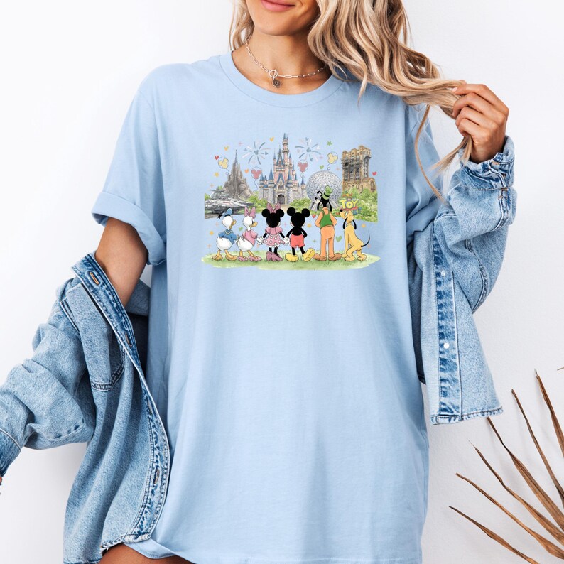 May include: Light blue t-shirt featuring a colorful illustration of Disney characters, including Mickey and Minnie Mouse, Donald and Daisy Duck, Goofy, and Pluto, in front of a castle and fireworks display. The shirt is a comfortable, casual style.