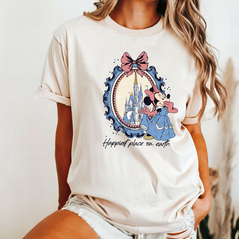 May include: A cream colored t-shirt with a cartoon image of Mickey Mouse in front of Cinderella's castle. The image is framed with a blue and gold ornate frame with a pink bow. The text "Happiest place on earth" is printed below the image.