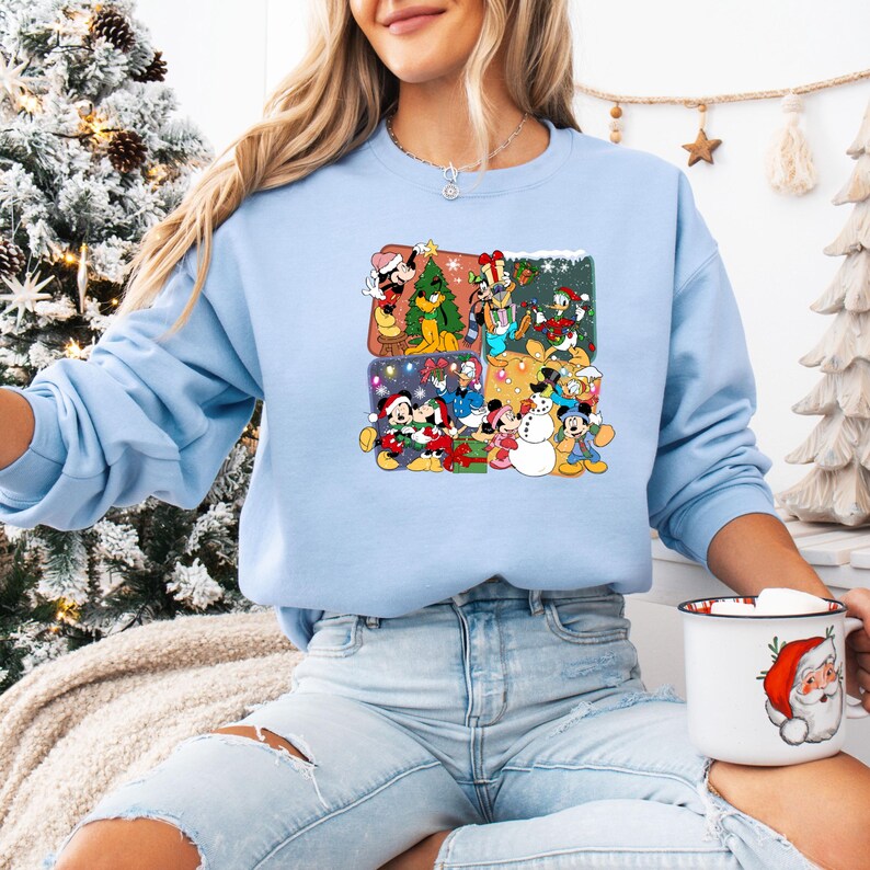 May include: A light blue sweatshirt featuring a colorful graphic of Disney characters celebrating Christmas. The graphic is divided into four squares, each depicting a different holiday scene with Mickey, Minnie, Donald, and Goofy. The sweatshirt is made of a soft material.