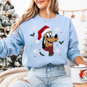 May include: A light blue sweatshirt featuring a cartoon dog wearing a Santa hat and a green and red scarf. The design includes snowflakes, holly, and gold sequins. The sweatshirt is part of a holiday-themed collection.