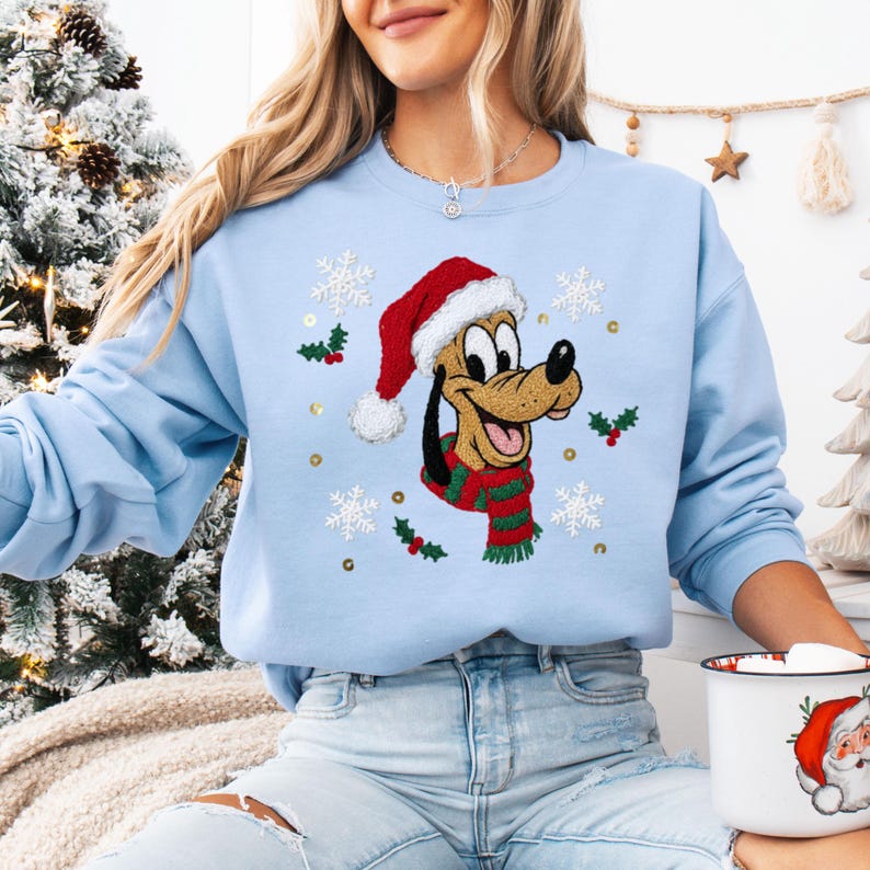 May include: A light blue sweatshirt featuring a cartoon dog wearing a Santa hat and a green and red scarf. The design includes snowflakes, holly, and gold sequins. The sweatshirt is part of a holiday-themed collection.