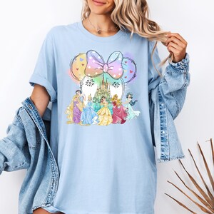 May include: A light blue t-shirt with a graphic of Disney princesses in front of Cinderella's castle. The princesses are wearing their signature dresses and the castle is surrounded by fireworks. The graphic is framed by a large pink bow with white polka dots.