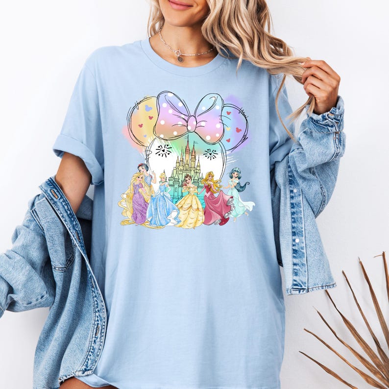 May include: A light blue t-shirt with a graphic of Disney princesses in front of Cinderella's castle. The princesses are wearing their signature dresses and the castle is surrounded by fireworks. The graphic is framed by a large pink bow with white polka dots.