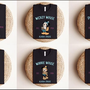 May include: Black t-shirts featuring Disney characters Pluto, Mickey Mouse, Donald Duck, Goofy, Minnie Mouse, and Daisy Duck. Each shirt has the character's name, the text "Alaska Cruise", and the year "1928" or "1924". The shirts are displayed on round woven mats.