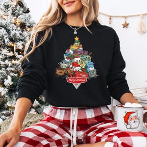 May include: Black sweatshirt featuring a Christmas tree design made of cartoon cars and characters. The tree is topped with a gold star and has the words "Merry Christmas" at the base. The sweatshirt is worn with red and white plaid pajama pants.